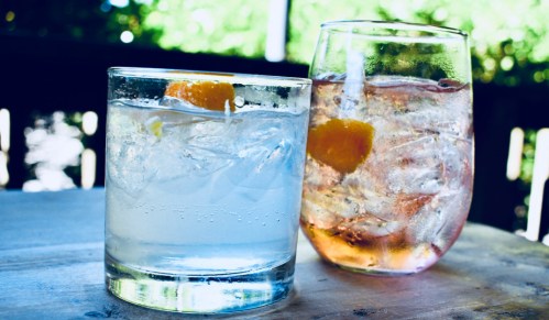 Two refreshing drinks on a wooden table: a clear cocktail with ice and a pink beverage with ice and orange slices.