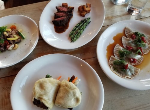 A table with four dishes: steak with asparagus, dumplings, a wrap, and a seafood pasta dish.