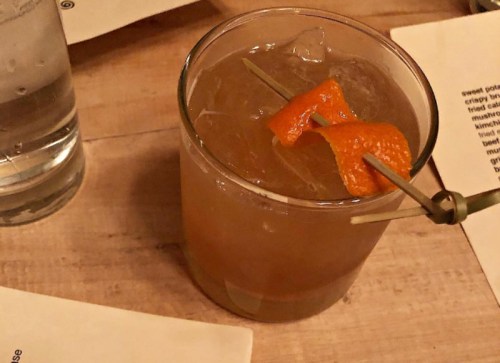 A cocktail in a glass with ice, garnished with an orange twist, on a wooden table next to a water glass and menus.