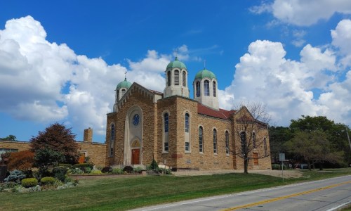 A stone church with green domes, surrounded by trees and a blue sky with fluffy clouds.