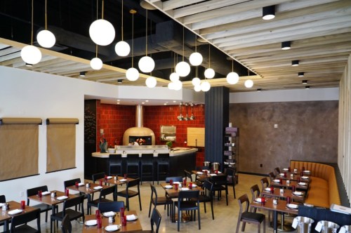 Modern restaurant interior with wooden beams, round pendant lights, and a pizza oven in the background. Tables set for dining.