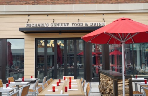 Exterior of Michael's Genuine Food & Drink, featuring outdoor seating, red umbrellas, and a wood stack.