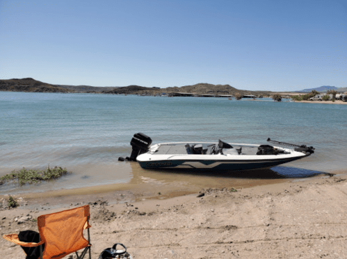 A small boat is anchored on a sandy shore by a calm lake, with a chair nearby and hills in the background.