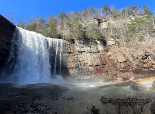 A majestic waterfall cascades over rocky cliffs, surrounded by trees, with a rainbow visible in the mist below.
