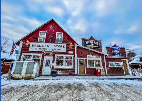A red wooden building with signs for Nagley's Store and West Rib Pub, surrounded by snow and a cloudy sky in Talkeetna, Alaska.
