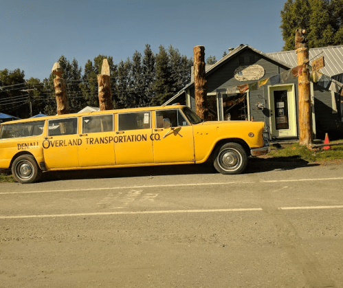 A vintage yellow limousine with "Denali Overland Transportation Co." signage parked outside a rustic building.