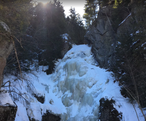 A frozen waterfall cascading down rocky cliffs, surrounded by snow-covered trees and a bright sun in the background.