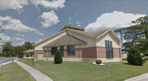 A modern church building with a cross on top, surrounded by grass and trees under a partly cloudy sky.