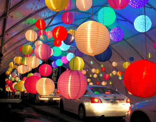 Colorful lanterns hang from a tent, illuminating a pathway where cars drive through a festive display.