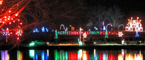 Colorful holiday lights display along a river, featuring a castle and "Merry Christmas from Holly Wild" sign.