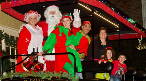 A festive group in holiday costumes, including Santa and an elf, smiles on a decorated float.