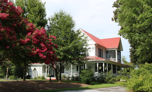 A charming two-story house with a red roof, surrounded by lush greenery and blooming pink flowers.