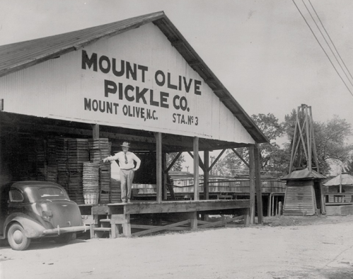 A man stands on the porch of the Mount Olive Pickle Co. building, with barrels and an old car nearby.