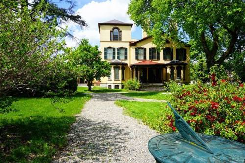 A large, historic house surrounded by greenery and colorful flowers, with a gravel path leading to the entrance.