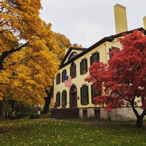 Historic house surrounded by vibrant autumn trees, featuring an American flag and colorful foliage.