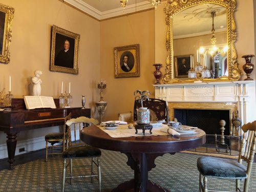 A vintage dining room with a round table, ornate mirror, portraits on the walls, and a grand piano in the corner.