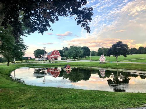 A red barn reflects in a calm pond, surrounded by green fields and trees under a colorful sky.