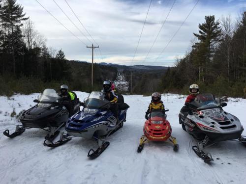 Four snowmobiles parked on a snowy trail, with riders in winter gear, surrounded by trees and mountains in the background.
