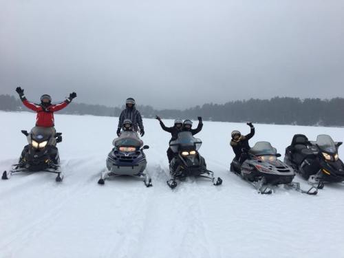 A group of five people on snowmobiles in a snowy landscape, smiling and raising their arms in excitement.
