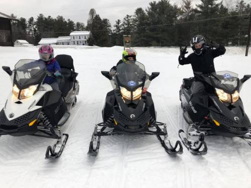 Three people on snowmobiles in a snowy landscape, smiling and posing for the camera. Trees and a building in the background.