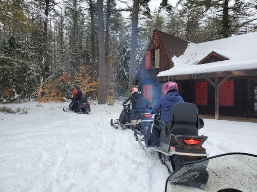 Three snowmobiles ride through a snowy landscape near a cabin surrounded by trees. Smoke rises from the cabin's chimney.