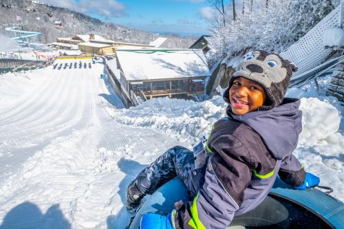 A smiling child in winter gear sits on a snow tube, ready to slide down a snowy hill on a sunny day.