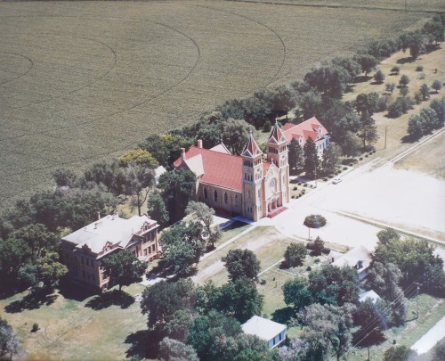 Aerial view of a historic building complex surrounded by trees and fields, featuring a large church and adjacent structures.