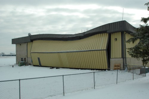 A building with a distorted, slanted exterior, surrounded by snow and a fence, under a cloudy sky.