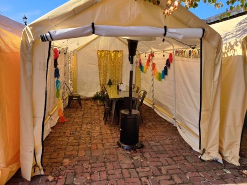 A cozy outdoor tent with a table, chairs, and a heater, decorated with colorful streamers and surrounded by brick flooring.