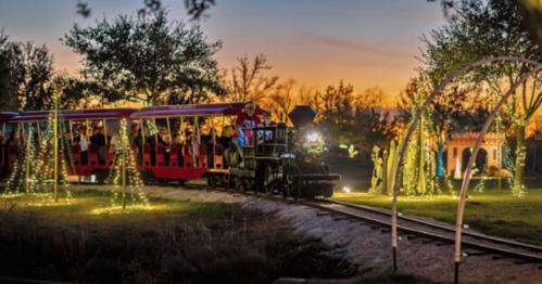 A festive train rides through a park at sunset, surrounded by twinkling lights and trees.