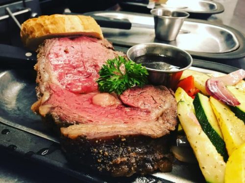 A juicy slice of prime rib served with vegetables and a side of sauce on a metal platter, with a piece of bread.