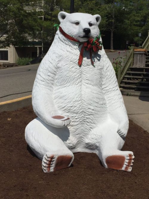 A large, white bear statue sits on the ground, wearing a festive red ribbon and surrounded by mulch.
