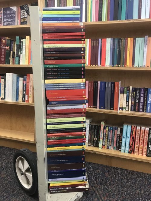A cart filled with stacked books in various colors, next to a bookshelf with more books in the background.