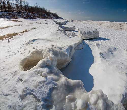 A snowy landscape with large ice formations along a shoreline under a clear blue sky.