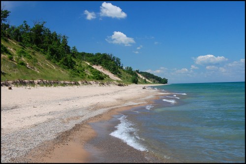 A serene beach scene with gentle waves, sandy shore, and lush green cliffs under a bright blue sky with fluffy clouds.