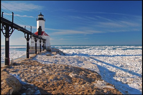 A lighthouse stands on a snowy shore, with ice-covered water and a clear blue sky in the background.