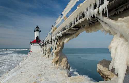 A frozen lighthouse on a pier, surrounded by ice and snow, with a calm lake in the background under a blue sky.