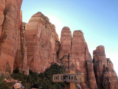 A sign reading "End of Trail" in front of towering red rock formations under a clear blue sky.