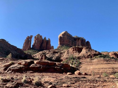 Red rock formations rise against a clear blue sky, showcasing layered textures and greenery at the base.