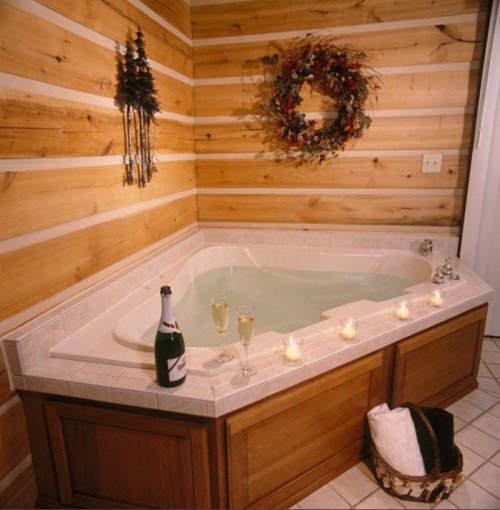 A cozy bathroom with a corner jacuzzi, candles, champagne, and a festive wreath on a wooden wall.