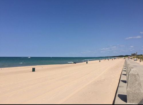 A sandy beach stretches along a calm blue ocean under a clear sky, with a walkway and distant people enjoying the scenery.