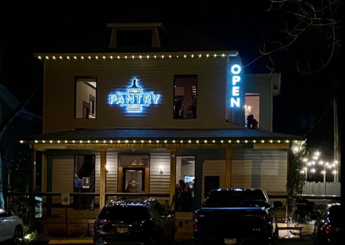 A cozy restaurant at night, illuminated with "Pantry" and "Open" signs, surrounded by string lights.