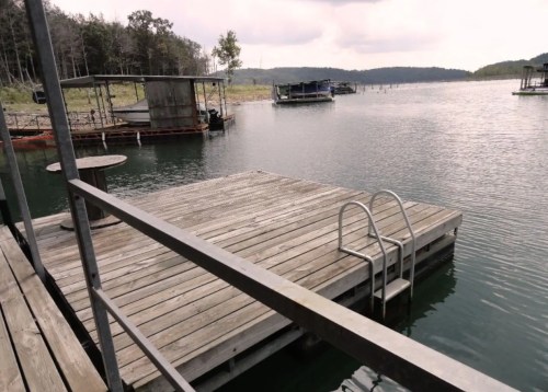 A wooden dock with a ladder extends into a calm lake, surrounded by trees and boats in the distance.
