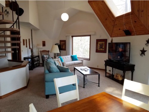 Cozy living room with a spiral staircase, large windows, and a TV, featuring a blue couch and wooden dining table.