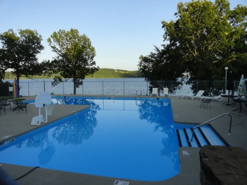 A serene swimming pool with clear blue water, surrounded by trees and overlooking a calm lake.
