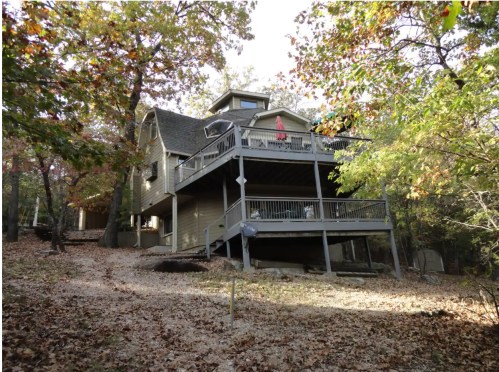 A two-story house surrounded by trees, featuring a large deck and a sloped yard covered in fallen leaves.