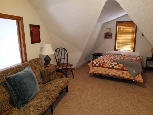 Cozy attic bedroom with a bed, quilt, armchair, and a lamp, featuring sloped ceilings and warm lighting.