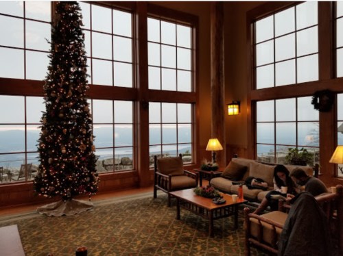 Cozy lodge interior with a Christmas tree, large windows, and a view of the mountains, featuring warm lighting and seating.