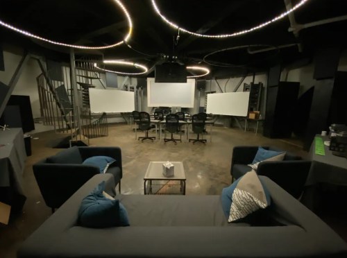 A modern meeting room with a dark aesthetic, featuring a sofa, chairs, and large screens on the walls.