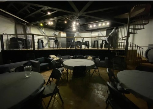 A dimly lit event space with round tables, chairs, and a spiral staircase leading to an upper level.